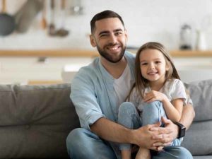 Portrait smiling father and little daughter hugging, looking at camera, sitting on cozy couch at home, happy young dad and cute adorable girl posing for family photo, enjoying leisure time together