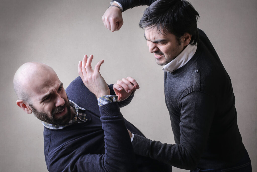 Two men are in a fight. The first man is committing battery, he is in the process of punching the second man, who has his arms up in a defensive posture to try to fend off the attack.